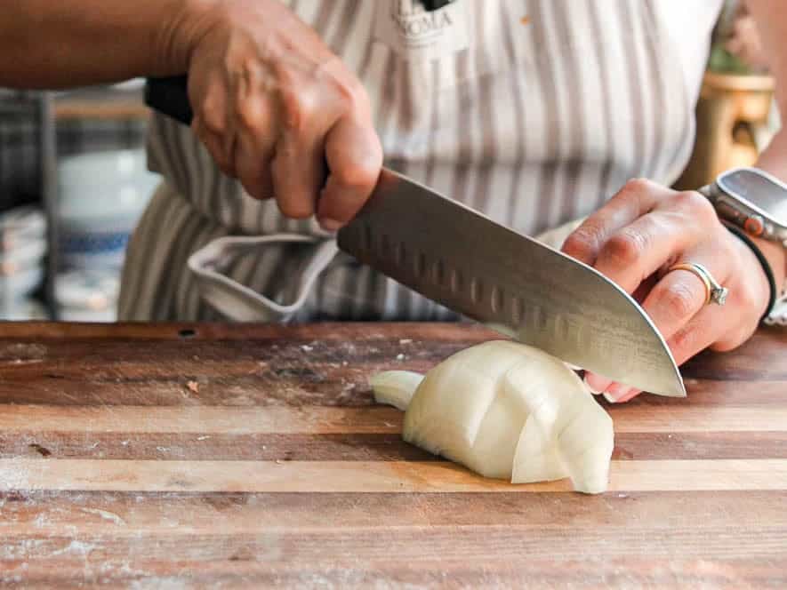 A person wearing a striped apron uses a large kitchen knife to slice a white onion on a wooden cutting board. Their left hand holds the onion steady while their right hand grips the knife. They are also wearing a watch on their left wrist.